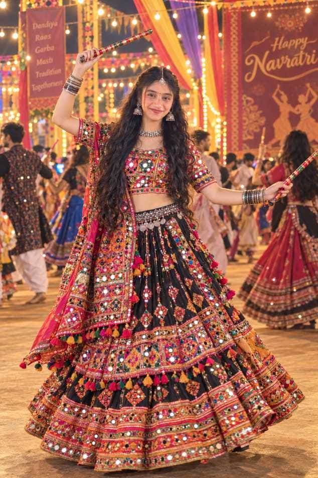 Smiling Indian girl with long curly hair in a black mirror-work lehenga holding a dandiya stick at a Garba venue