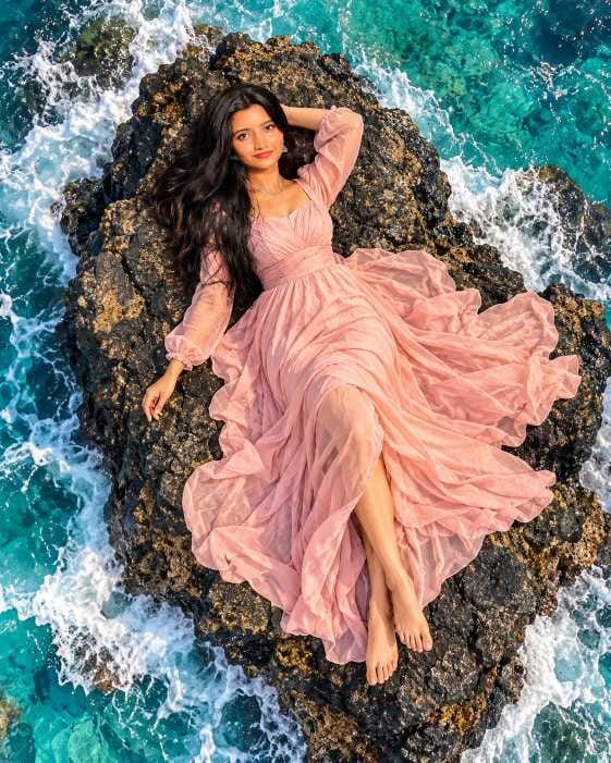 Drone view AI photo prompt for girls showing a girl in a flowing pink dress lying on a rock in the sea