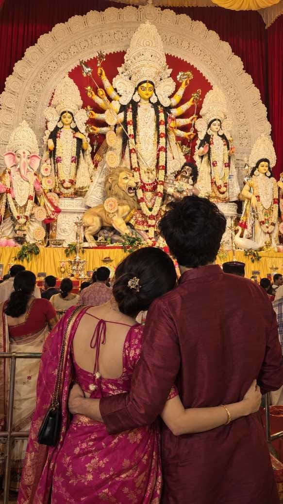 Couple seen from behind hugging at the waist before a Durga idol stage, she in a pink saree, he in a maroon kurta