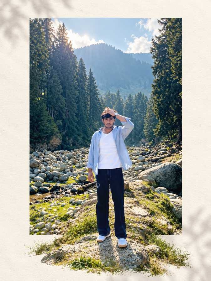 Young man with a short beard standing on a grassy riverbed with tall pine trees and a misty mountain behind him